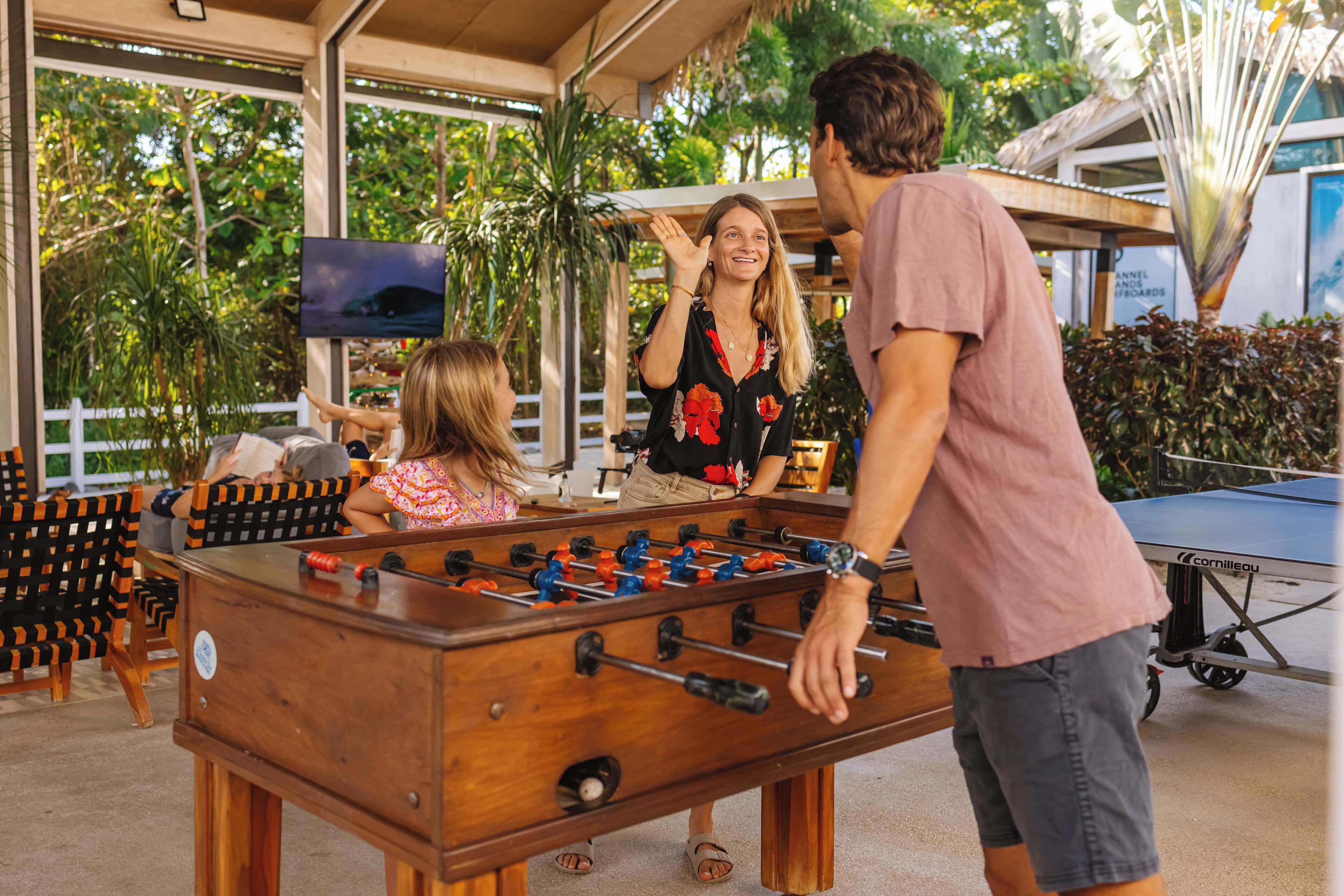 A foosball table in a covered outdoor area with chairs and a television screen.