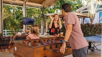 A foosball table in a covered outdoor area with chairs and a television screen.