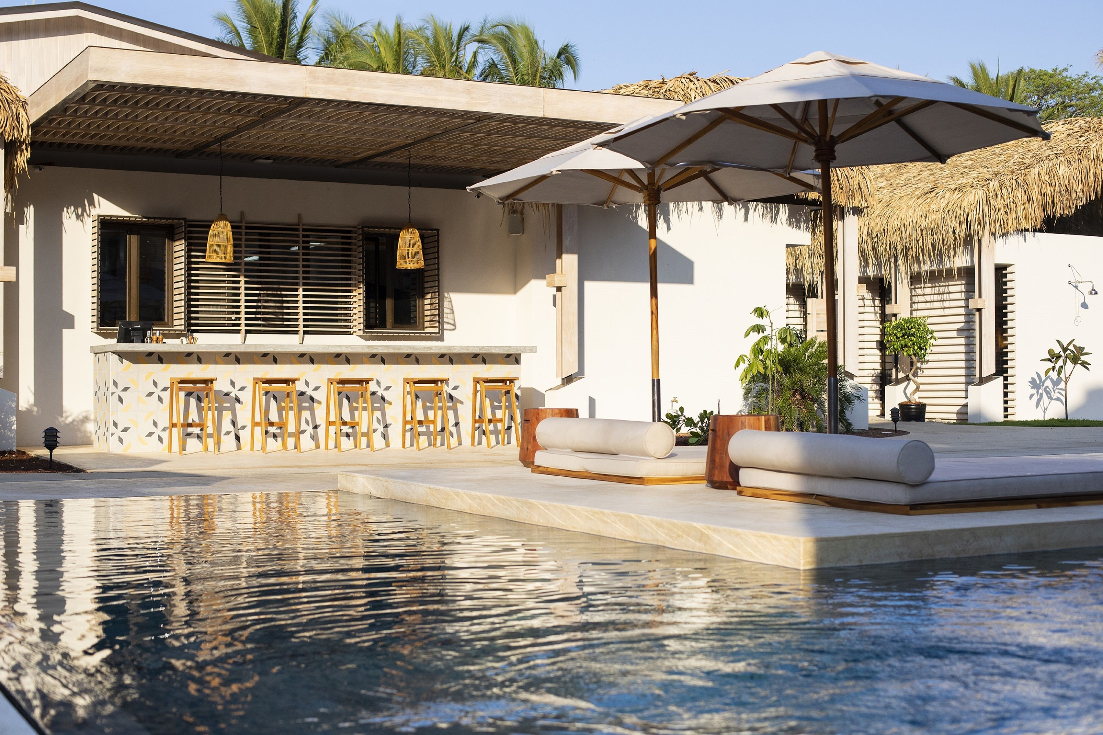 A pool area with a bar and seating, surrounded by a white building with a thatched roof and palm trees.