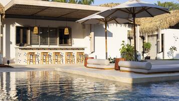 A pool area with a bar and seating, surrounded by a white building with a thatched roof and palm trees.