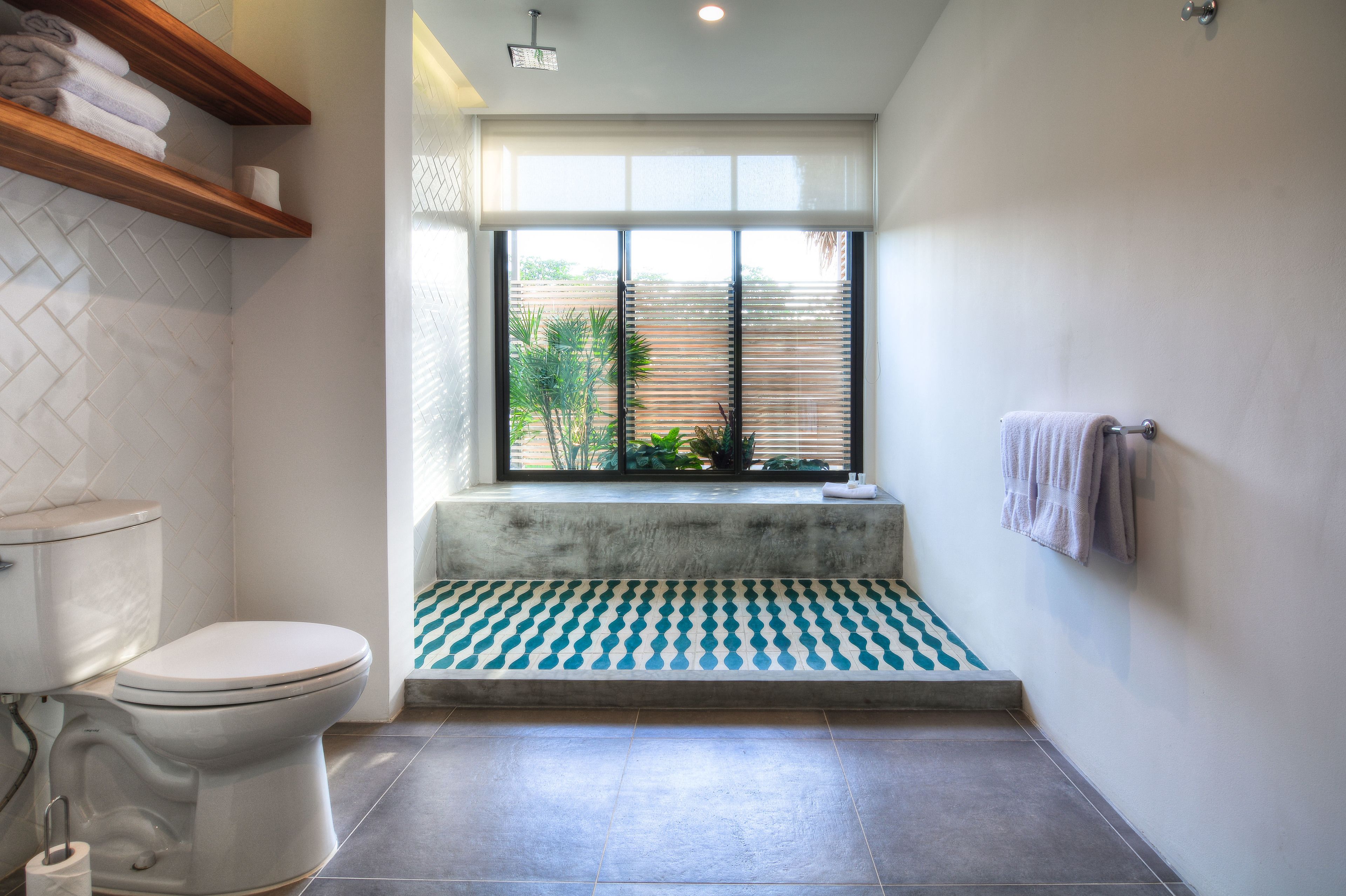 Une salle de bain comprenant des toilettes, une fenêtre avec des stores, un porte-serviettes et un tapis à motifs.
