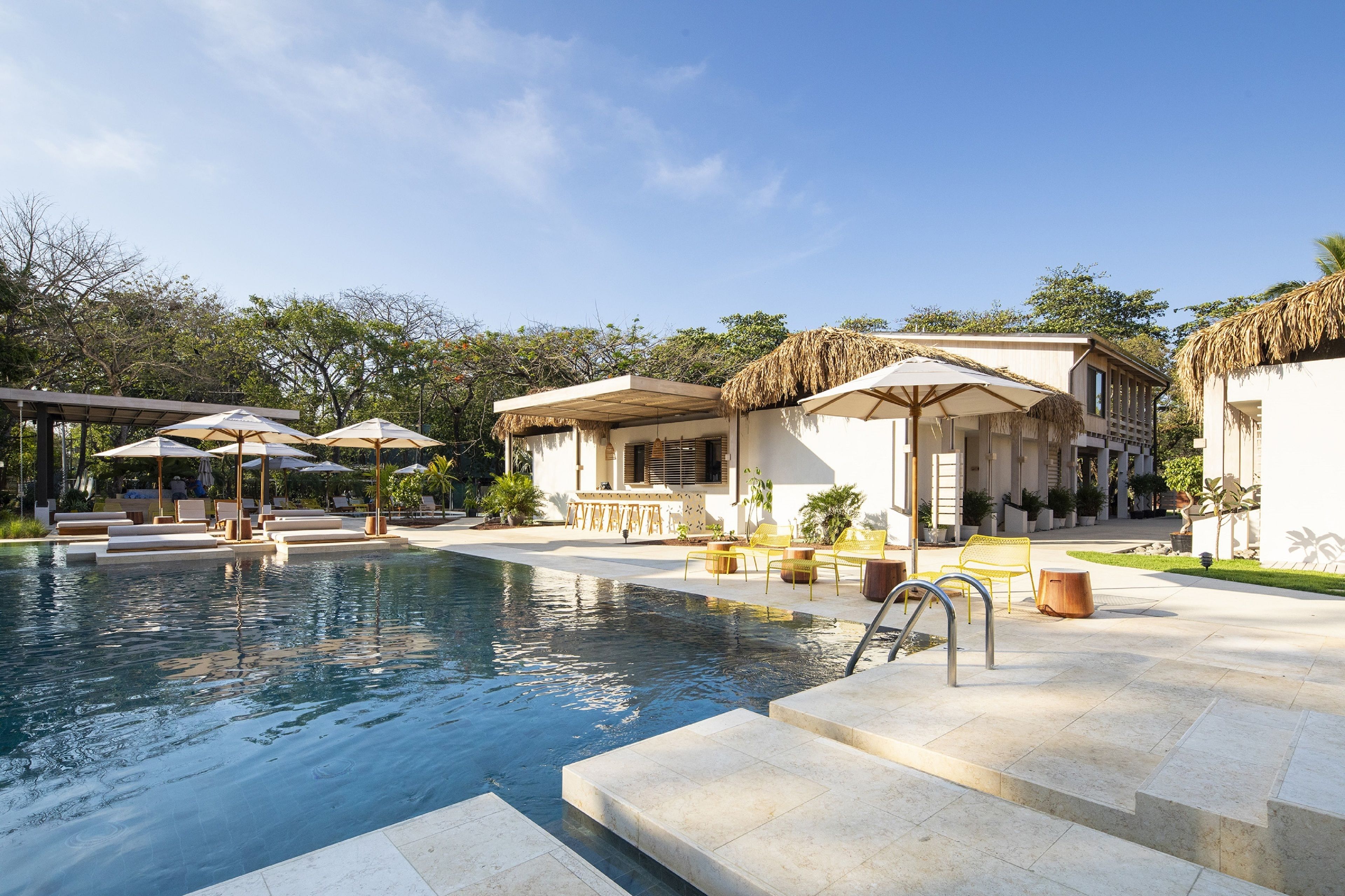 A pool area with lounge chairs and umbrellas, a bar, and a building with a thatched roof.