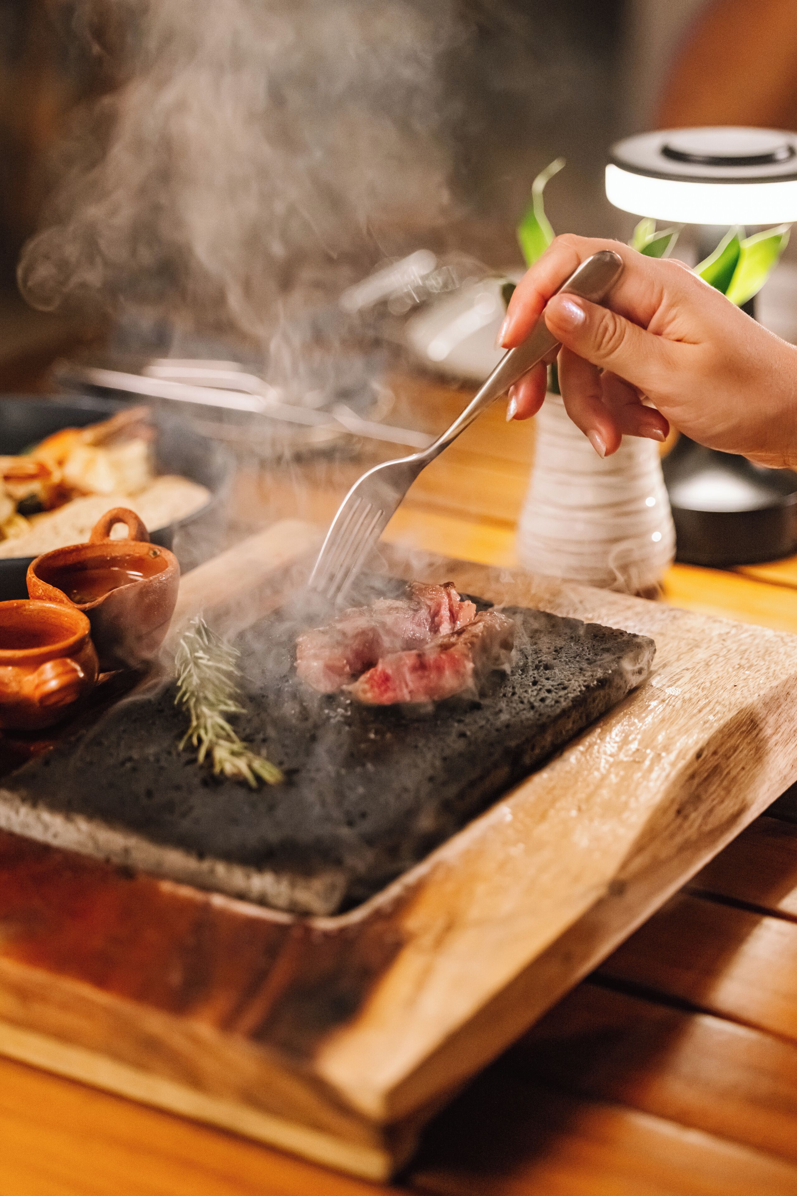 A person grilling meat on a charcoal grill with a fork, surrounded by cooking utensils and a wooden cutting board.