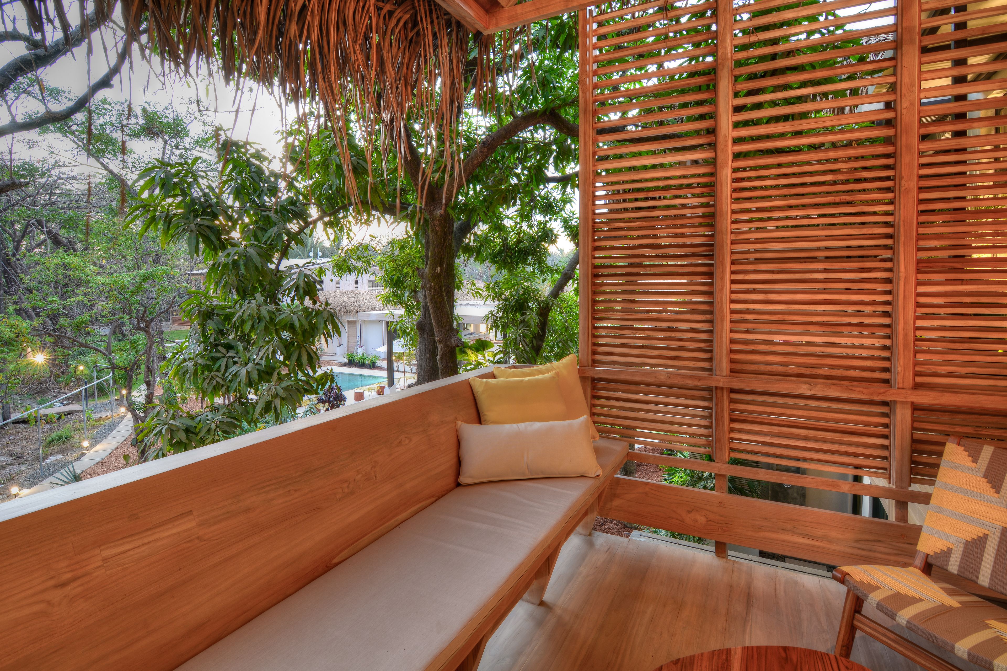 A wooden bench with cushions, a wooden table, and a wooden slatted wall with a view of trees and a building in the background.
