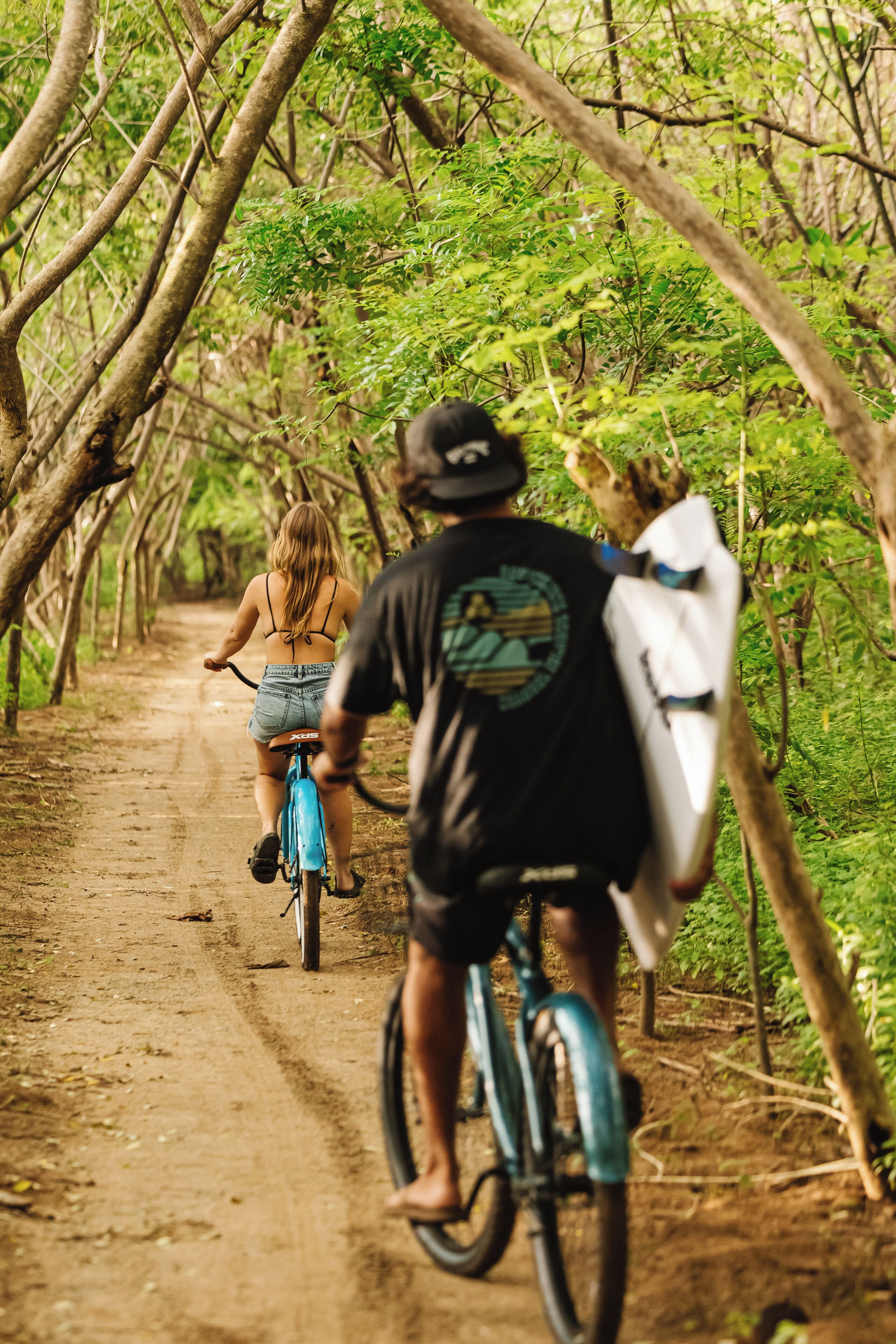 Un sendero de tierra bordeado por árboles, con una persona montando en bicicleta y otra llevando una tabla de surf.