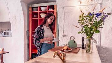 A woman in a floral shirt holding a cup, standing in a kitchen with a wooden table, a kettle, and a vase of flowers.