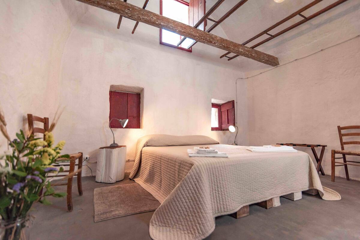 Un dormitorio con una cama, dos sillas, una mesita, un ventanal y techo con vigas de madera.