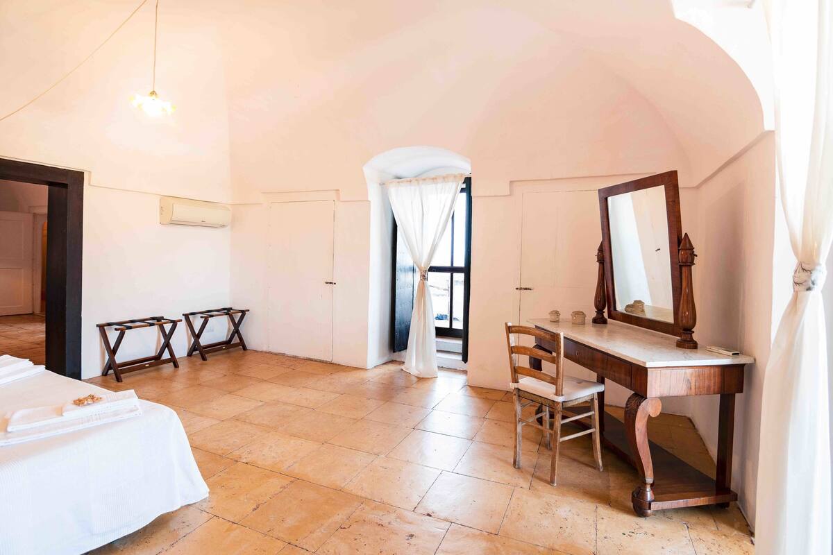 Una habitación de hotel con cama, un escritorio con espejo y una silla. Hay una puerta que da a otra habitación y una ventana con cortinas.
