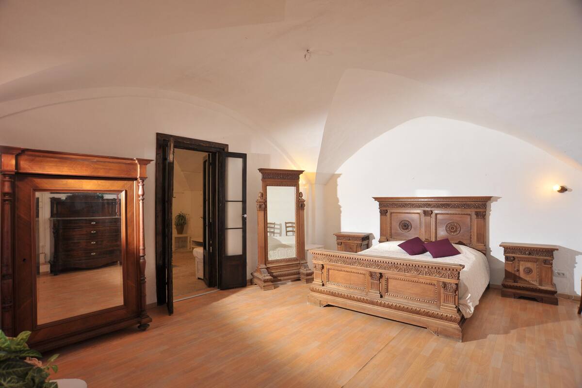 Un dormitorio con una cama grande, un aparador de madera, un espejo y una puerta que da a otra habitación.