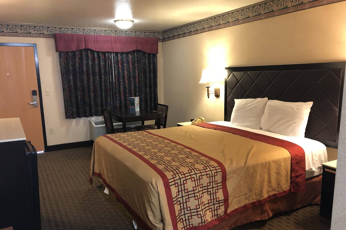 A hotel room with a large bed, a desk with a chair, a telephone, and a wall-mounted lamp.