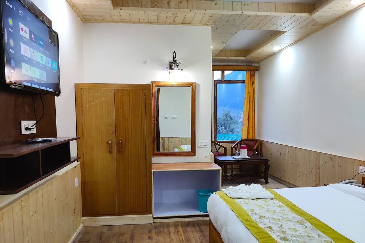 A bedroom with a wooden wardrobe, a bedside table, a bed with a green and white bedspread, a TV mounted on the wall, and a window with a view of the outside.