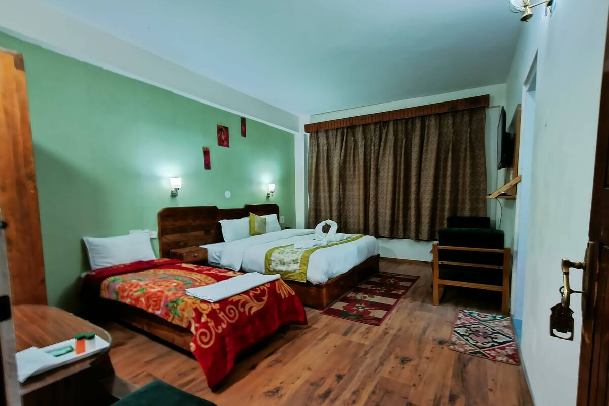 A hotel room with a wooden floor, two beds with red and floral bedding, a green armchair, a wooden desk, and a TV on the wall.