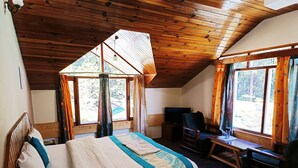 A cozy bedroom with a wooden ceiling, a bed with a blue and white bedspread, a desk with a chair, and large windows offering a view of the outdoors. - CS Shhandella Hotel (Manali)