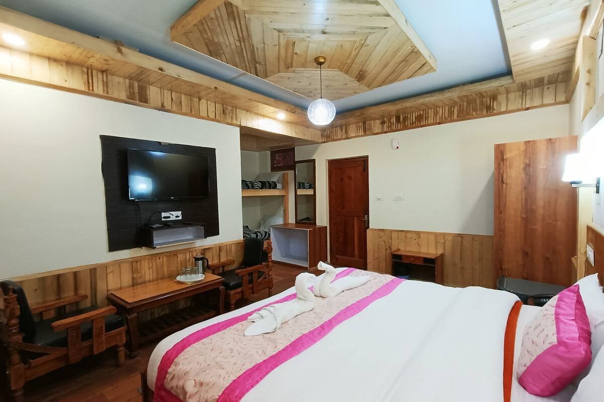 A bedroom with a wooden ceiling, a bed with a pink and white bedspread, a television mounted on the wall, and a wooden desk with a chair.