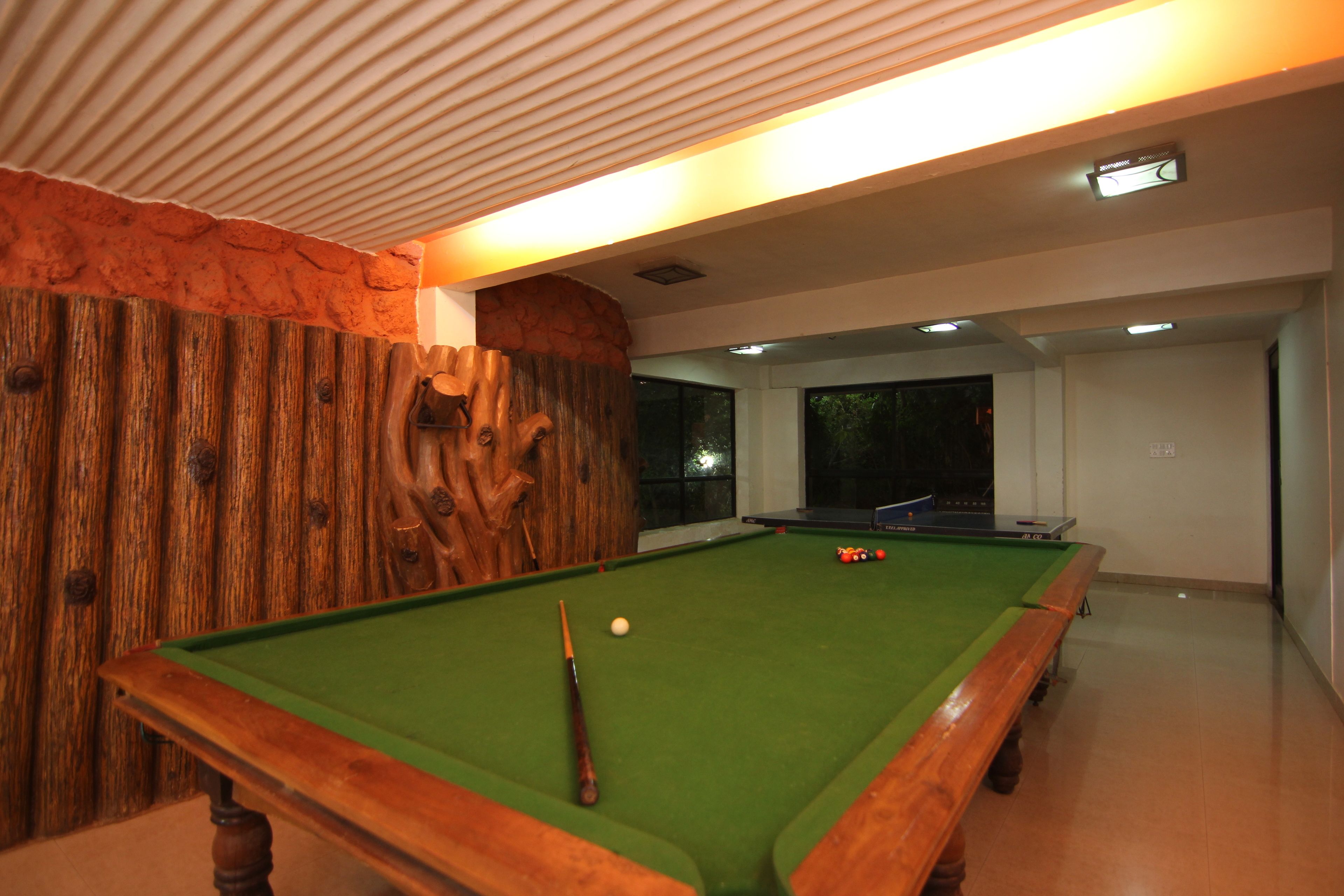 A pool table with a wooden frame and green felt, a cue stick, and billiard balls in a room with a wooden accent wall and a window in the background.