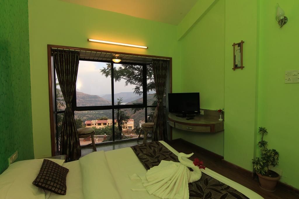 Habitación de hotel con cama, televisor sobre un mueble, ventana con cortinas y vistas al exterior.