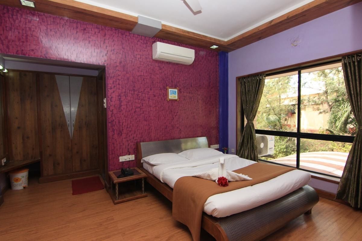 A hotel room with a bed, wooden wardrobe, air conditioning unit, and a window with curtains.