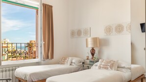 A hotel room with two beds, a window with a view of buildings and palm trees, a table with a lamp, and wall art. - Guest House Maison 6 (Rome)