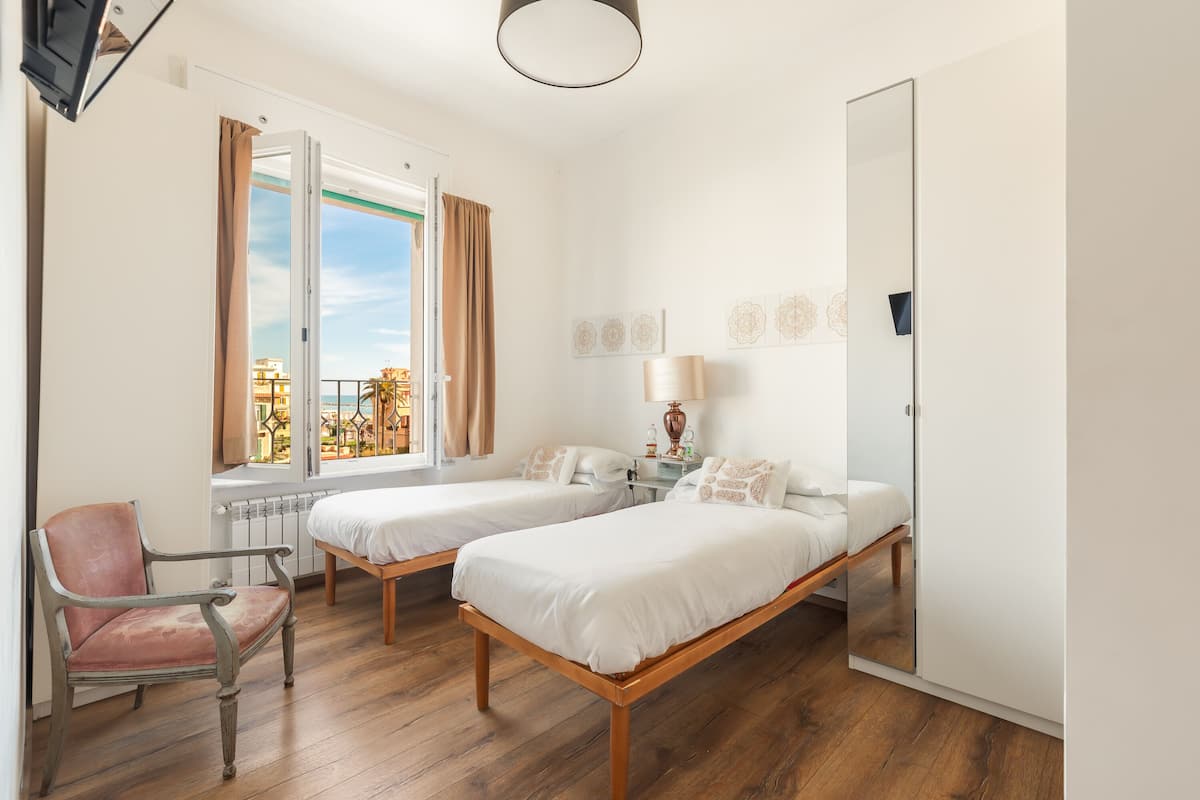 A hotel room with two single beds, a chair, a bedside table with a lamp, a mirror, and a window with curtains.