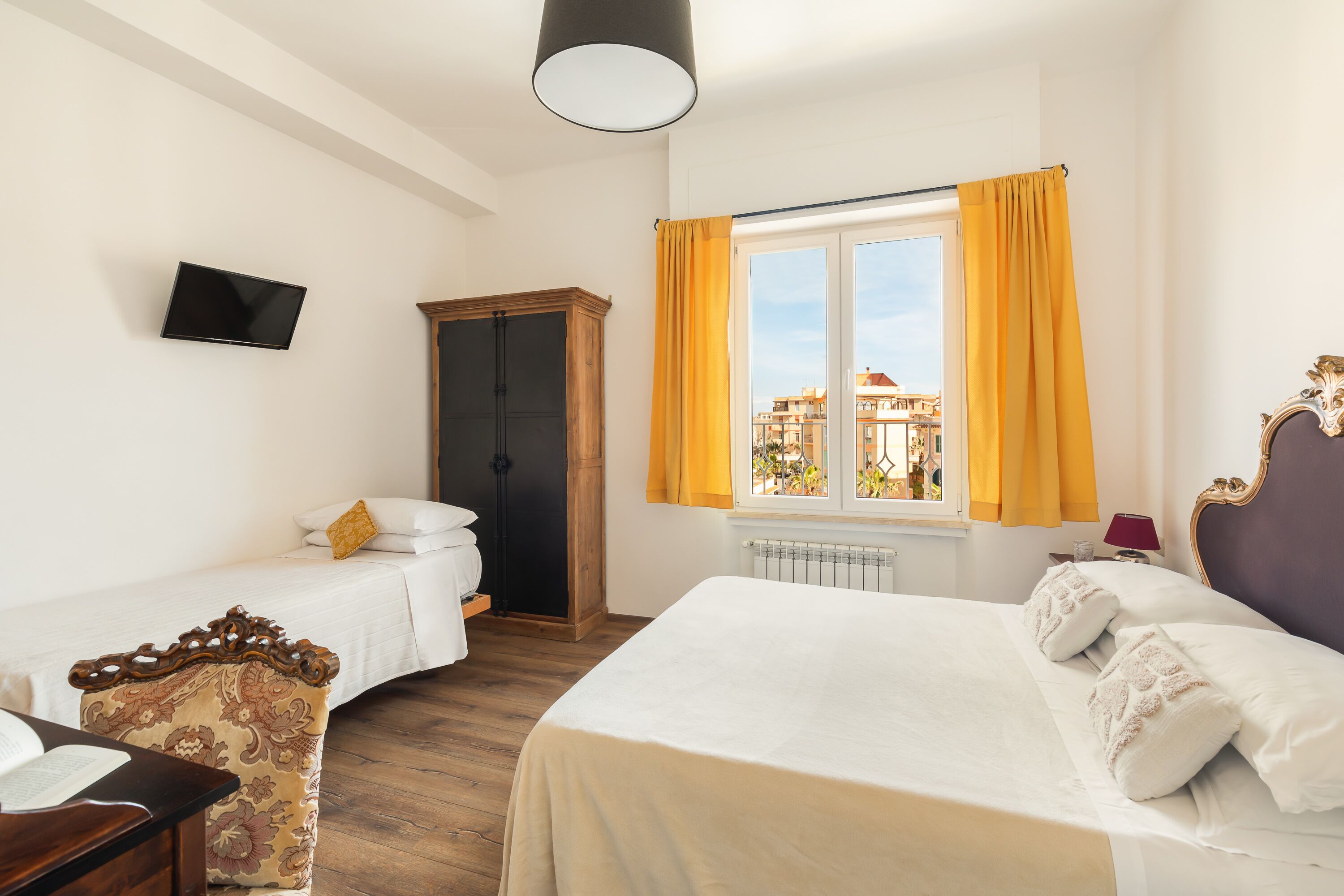 A hotel room with two beds, a TV, a wooden wardrobe, and a window with curtains.