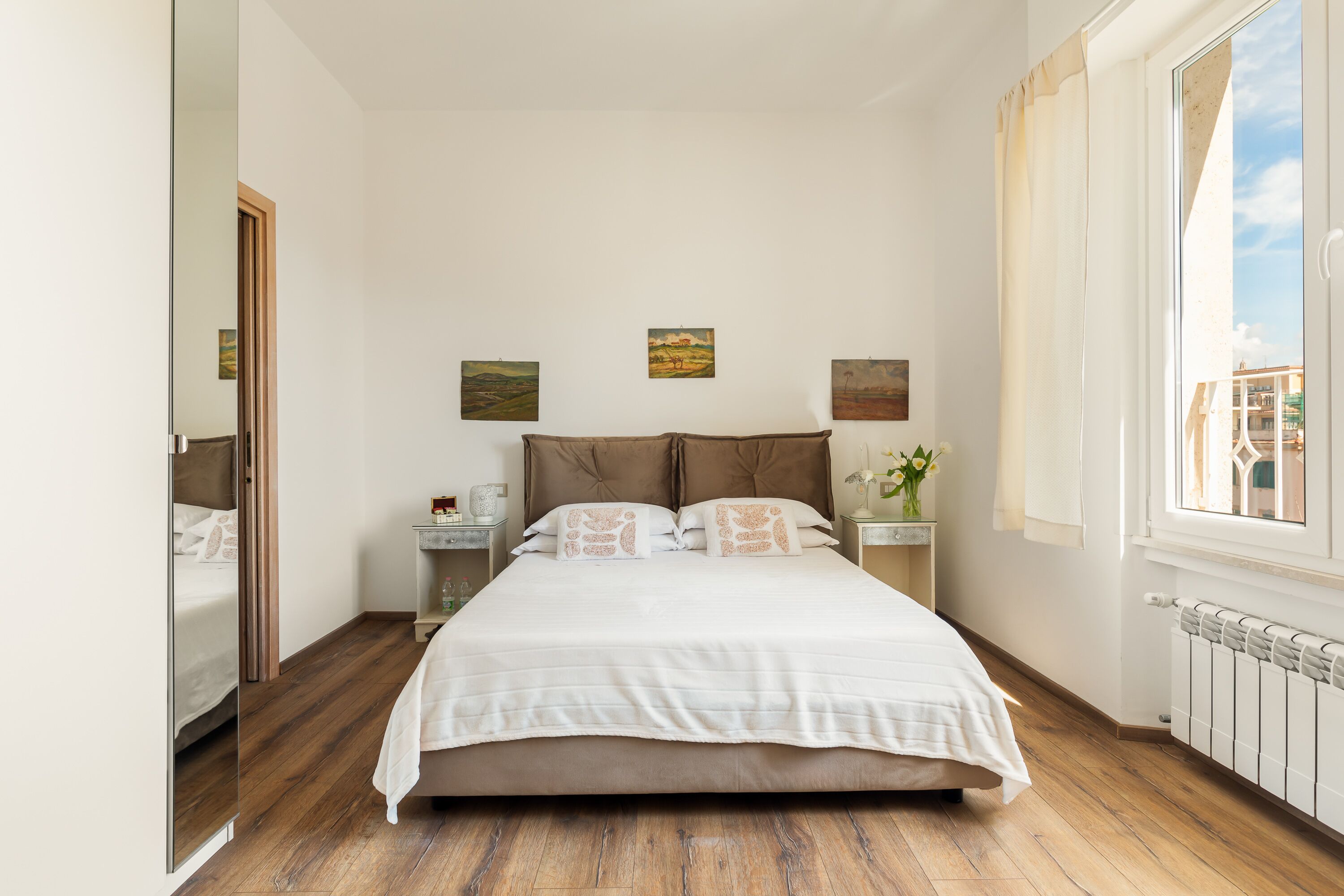 A bedroom with a large bed, wooden flooring, a window with curtains, and three paintings on the wall.