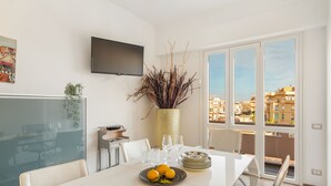 A dining area with a white table, chairs, a plate of fruit, a vase with plants, and a TV on the wall. - Guest House Maison 6 (Rome)