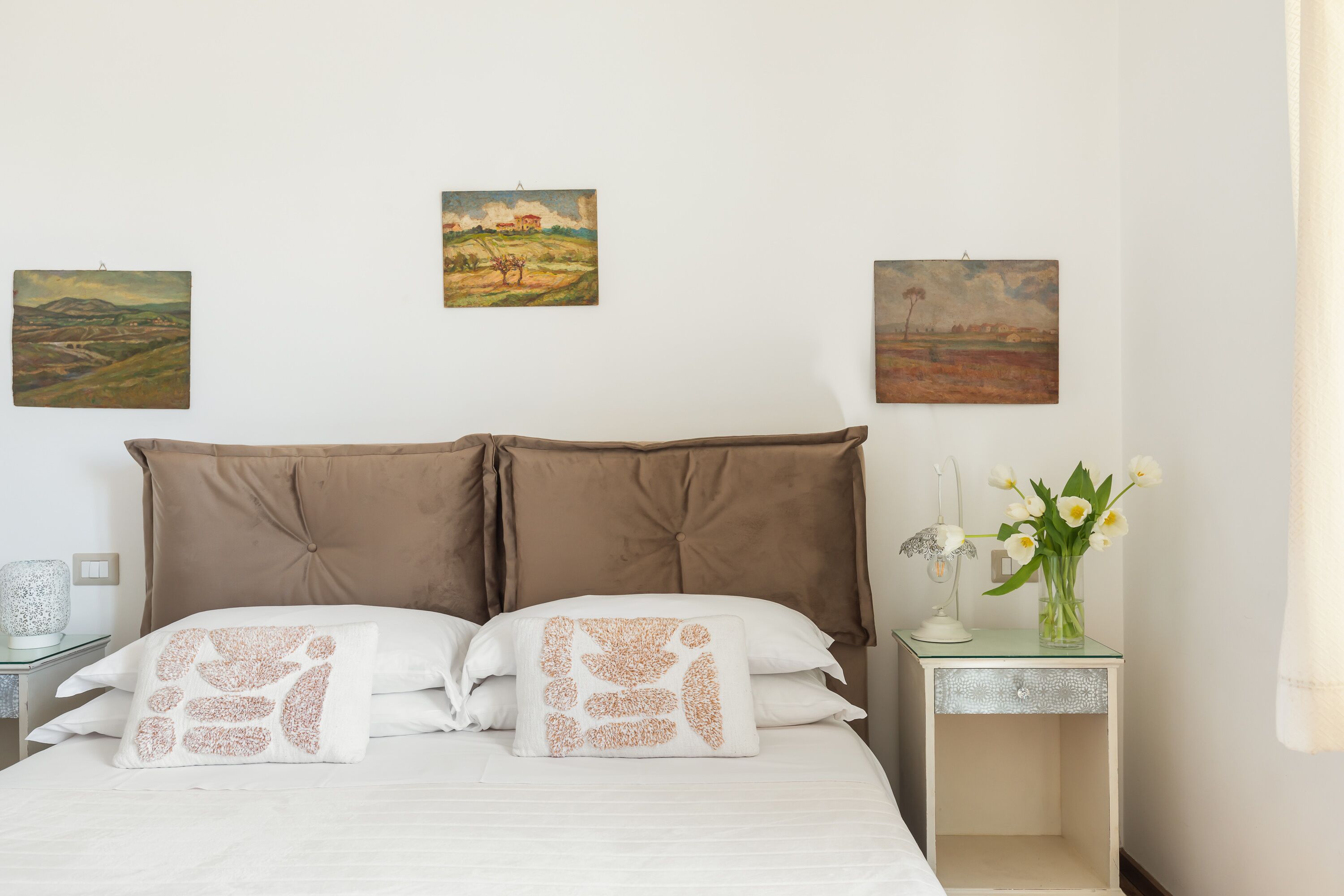 A bedroom with a bed, two pillows, a nightstand with a lamp, and three paintings on the wall.