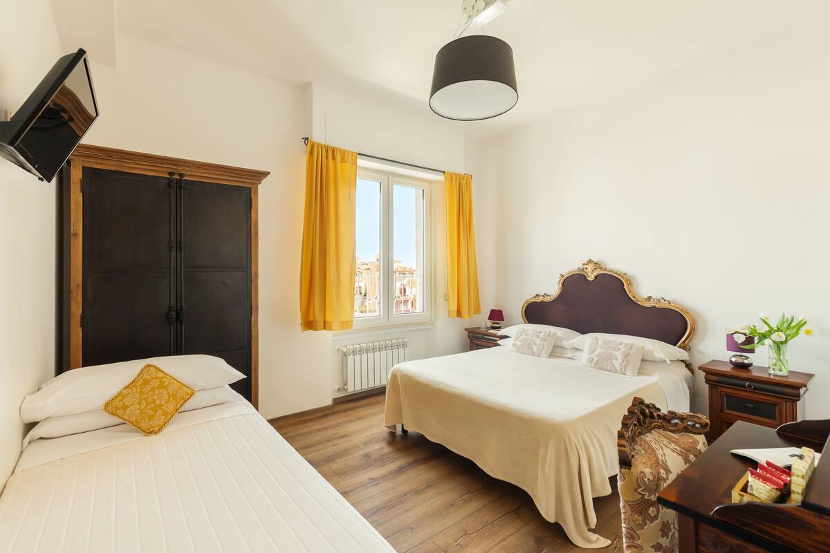 A bedroom with two beds, a wooden floor, a radiator, a window with yellow curtains, a bedside table with a lamp, and a nightstand with a vase of flowers.