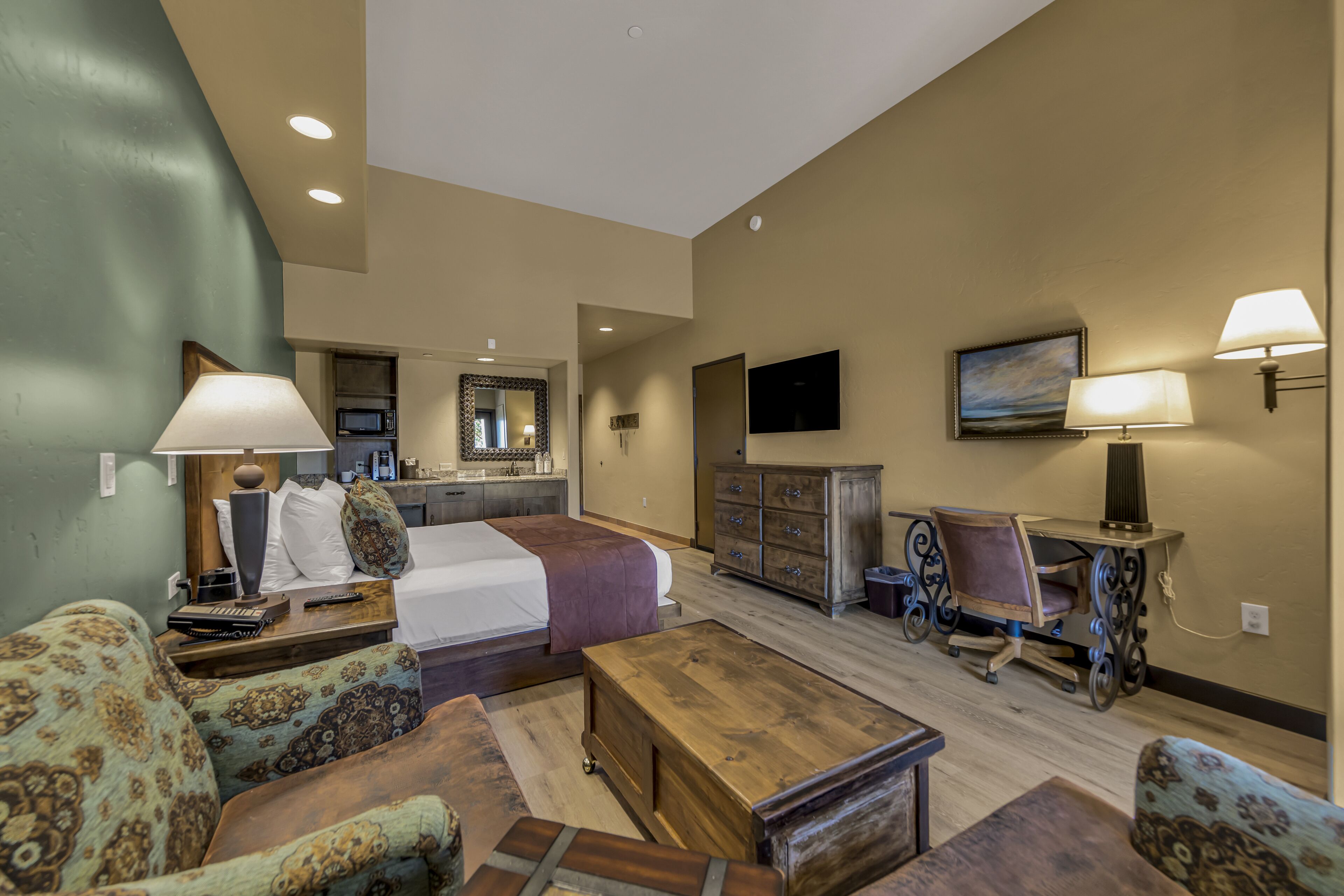 A spacious living room with a large bed, a wooden coffee table, patterned sofas, a dining table with chairs, a TV mounted on the wall, and a kitchen area in the background.