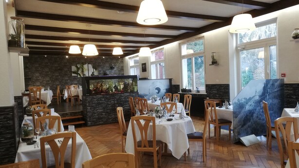 Un restaurante con suelos de madera, manteles blancos y sillas de madera. Se observan luces colgantes y un acuario grande al fondo.