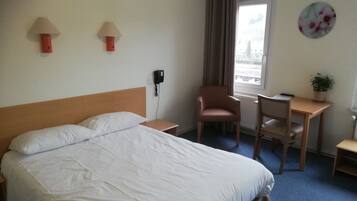 A hotel room with a bed, a desk, a chair, a phone, a window with curtains, and a wall decoration.