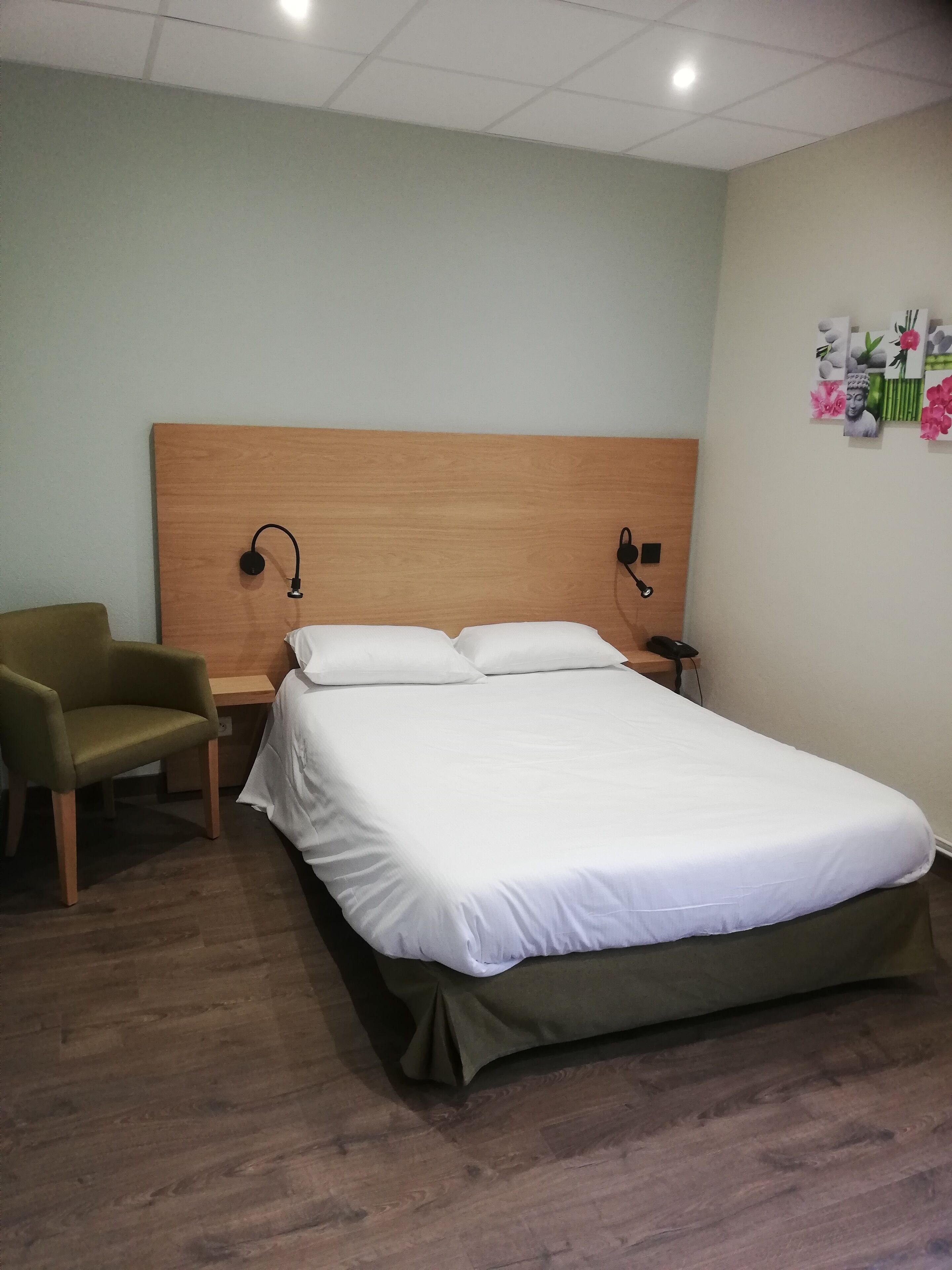 A hotel room with a single bed, a wooden headboard, two bedside lamps, a green armchair, and a nightstand with a phone.