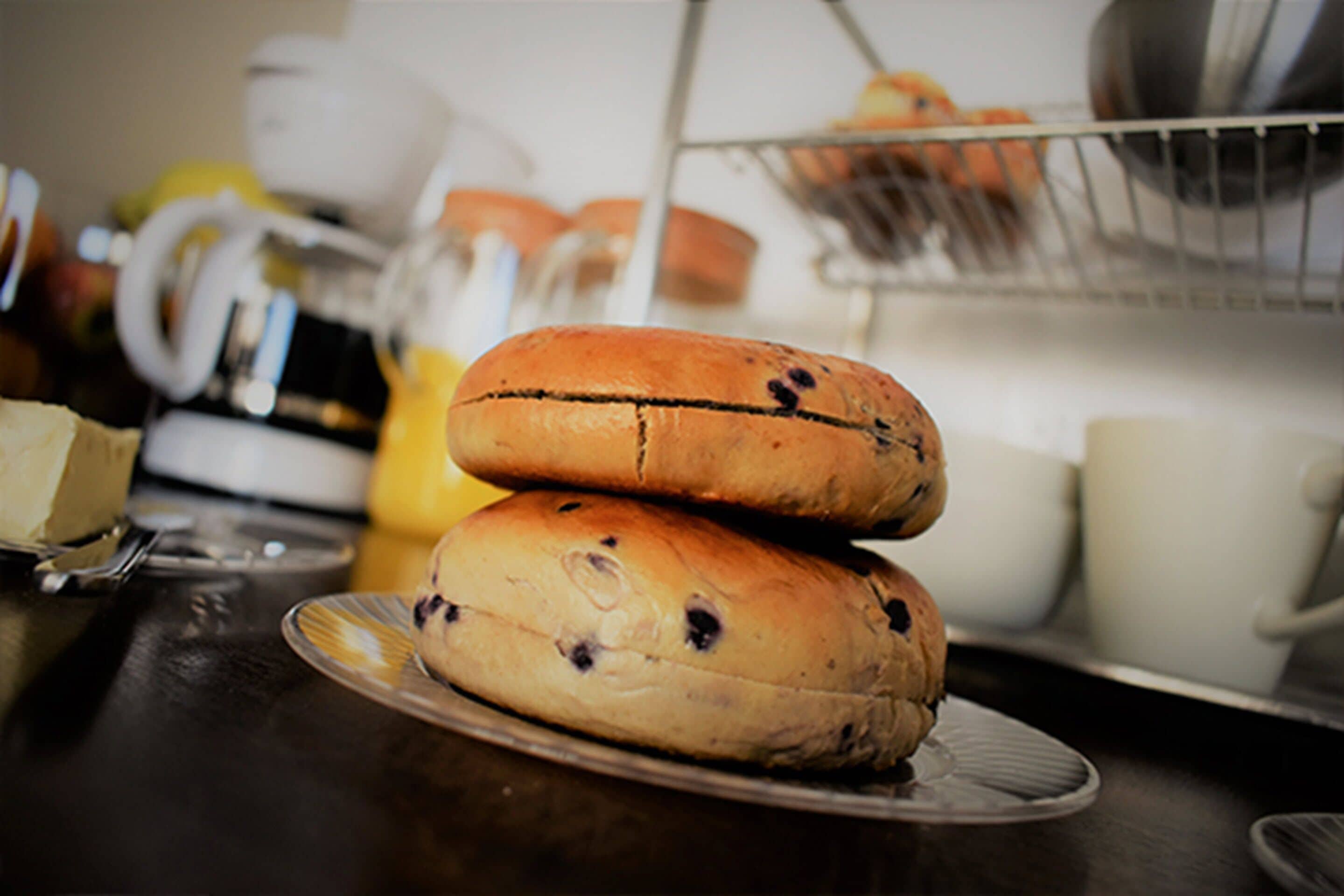 A kitchen counter with two stacked bread rolls on a plate, a coffee maker, and a fruit basket in the background.