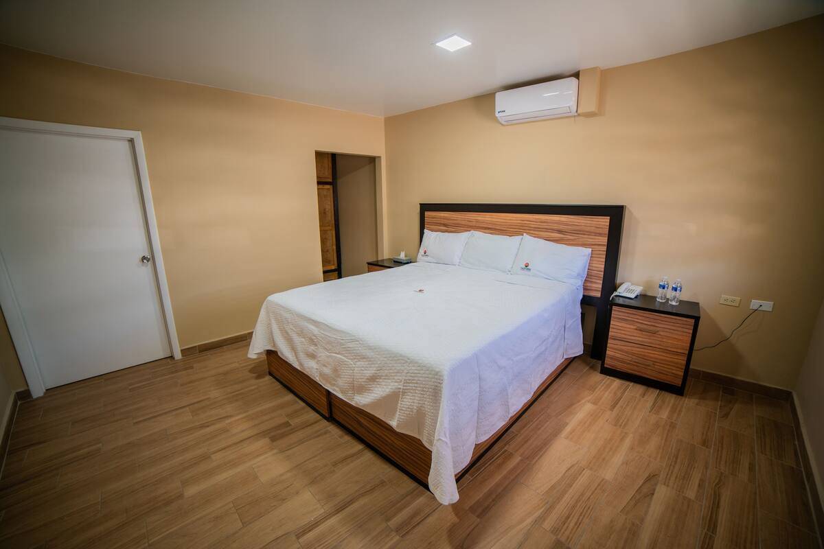A bedroom with a wooden bed, white bedding, a nightstand with a phone and water bottles, and an air conditioning unit on the wall.