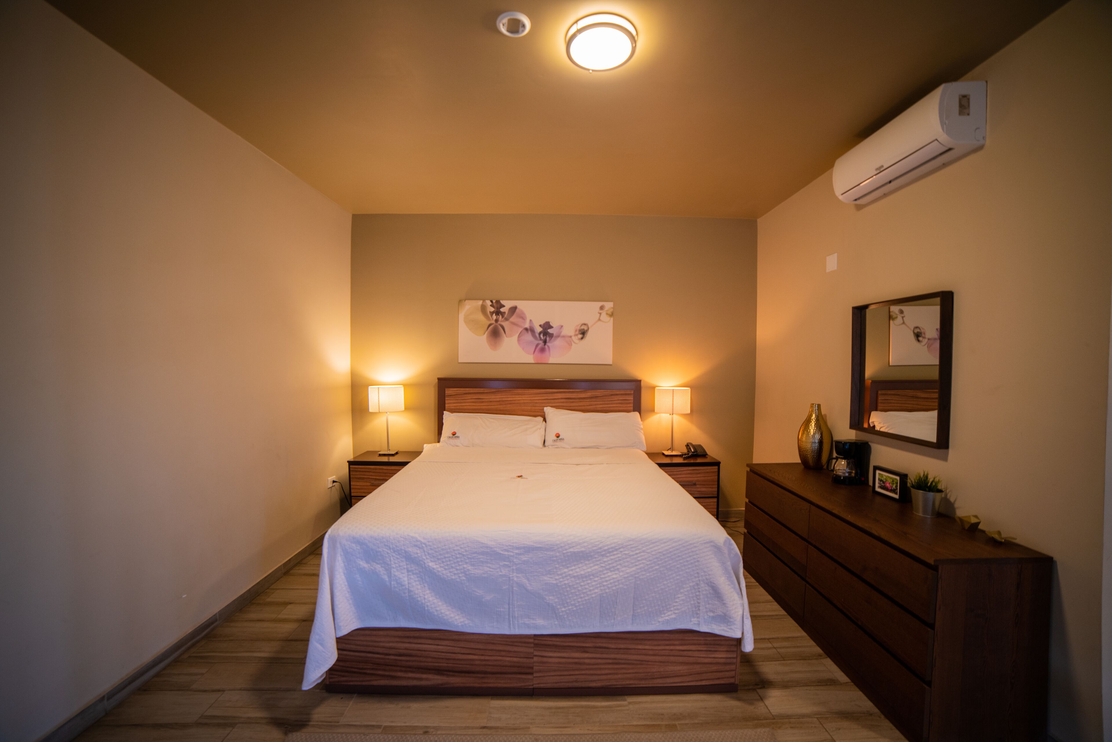 A neatly made bed with a wooden headboard, two bedside lamps, a nightstand, a dresser, a wall-mounted air conditioner, and a framed picture above the bed.