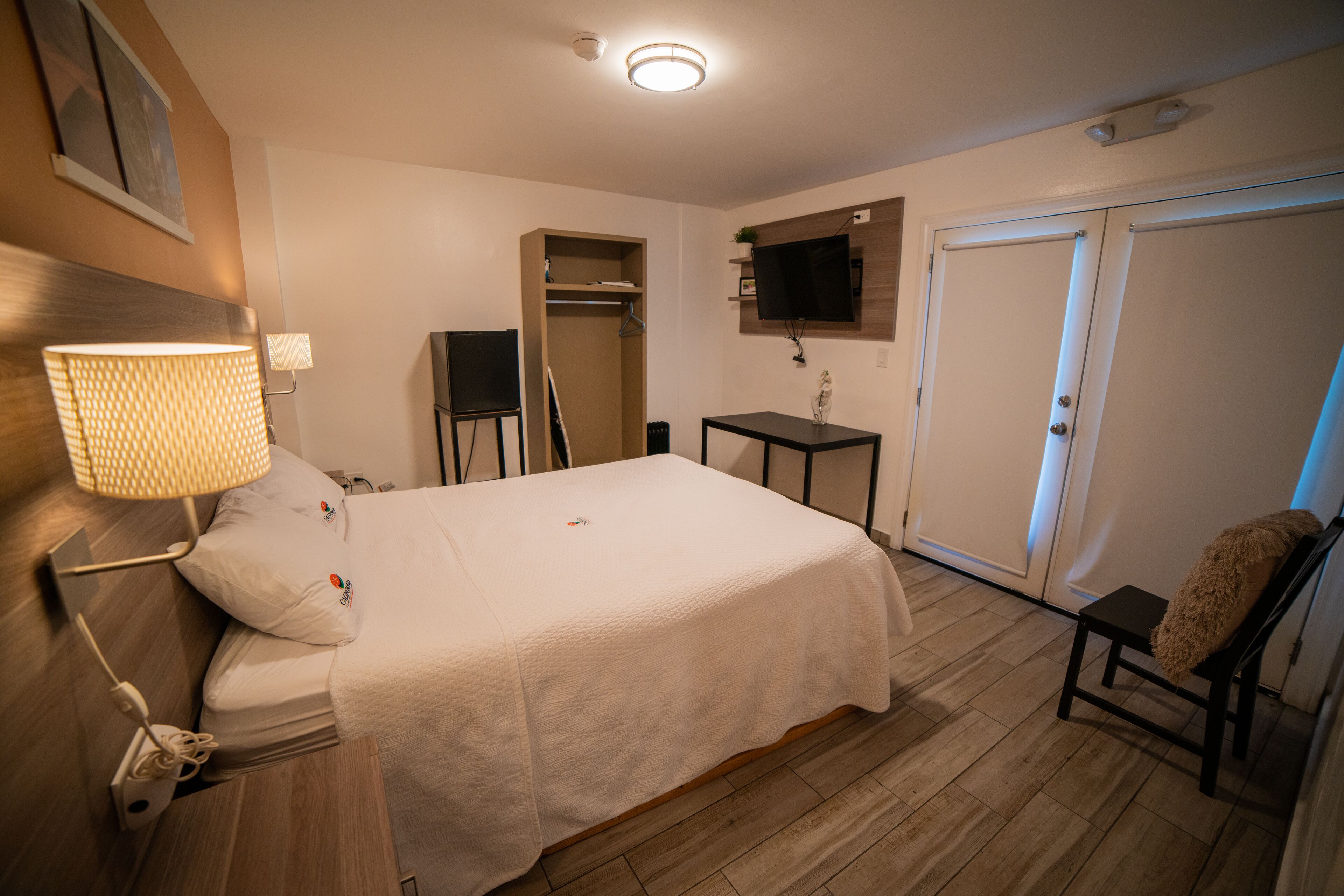 Una habitación de hotel con cama, mesita de noche, lámpara, televisor y armario.