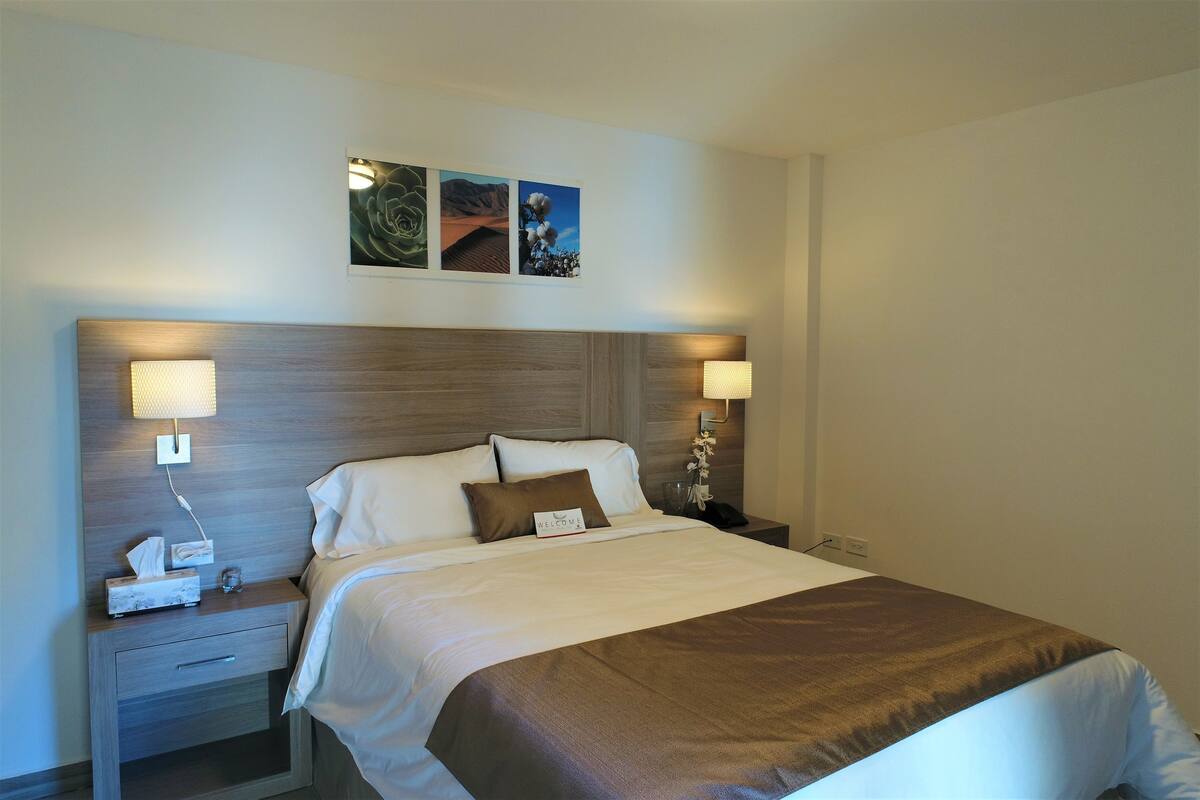 A neatly made bed with a brown accent throw, a bedside lamp, and a nightstand with a phone and a tissue box.