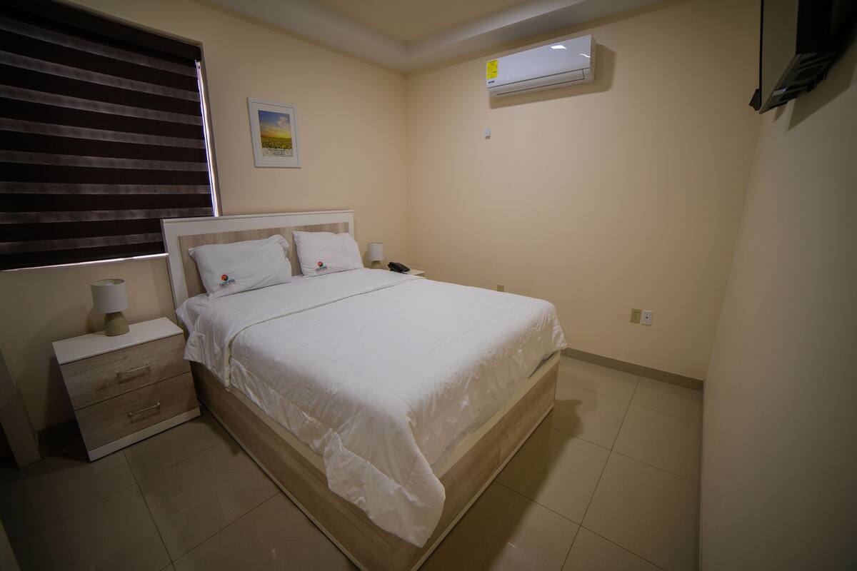 A bedroom with a bed, bedside tables, a nightstand, an air conditioner, and a wall-mounted TV.