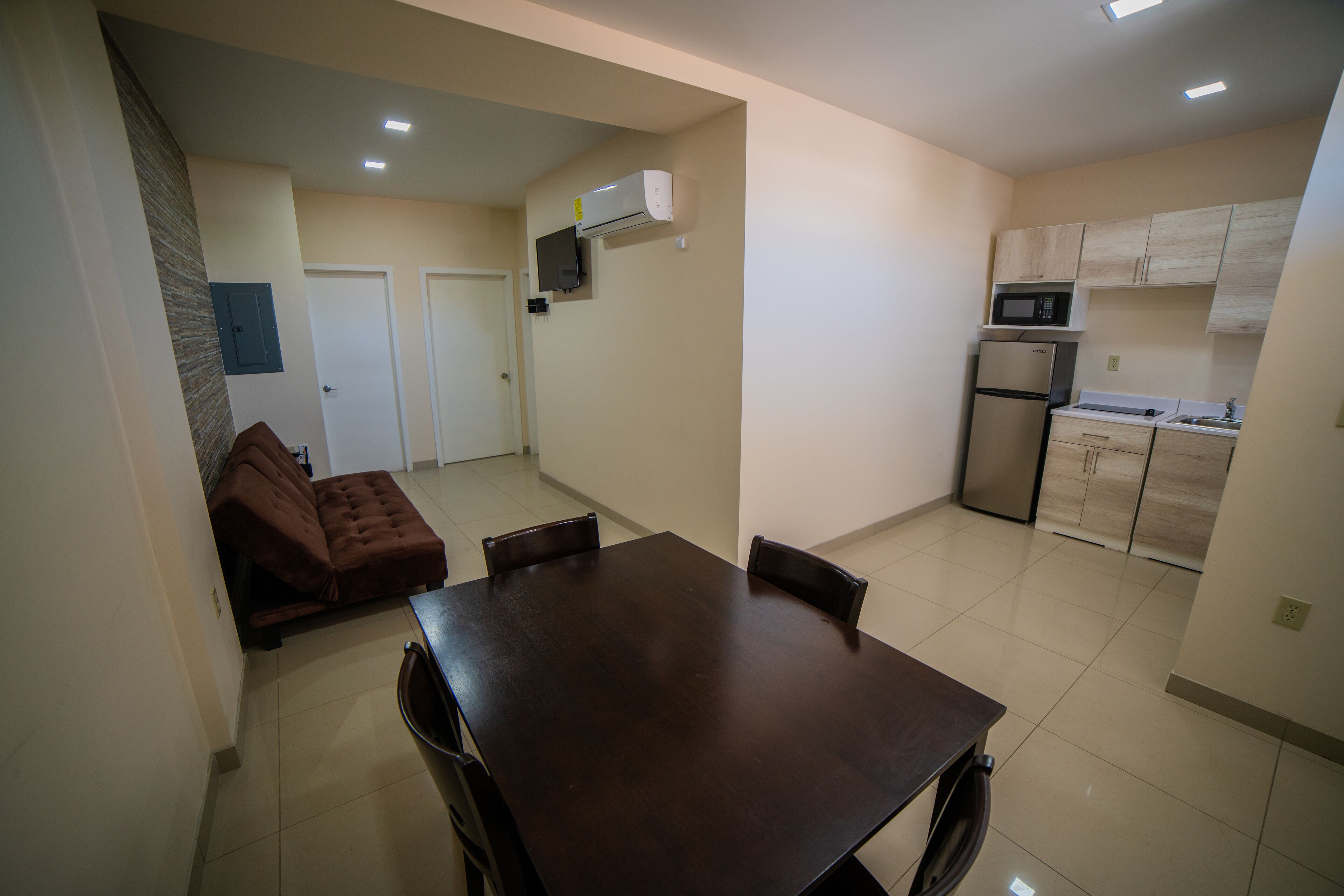 A compact kitchen with a refrigerator, microwave, and air conditioning unit. There is a dining table with chairs and a sofa in the adjacent living area.