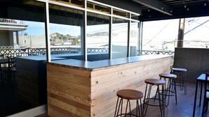 Een bar met een houten toonbank en krukken, grote ramen en uitzicht op gebouwen en heuvels.