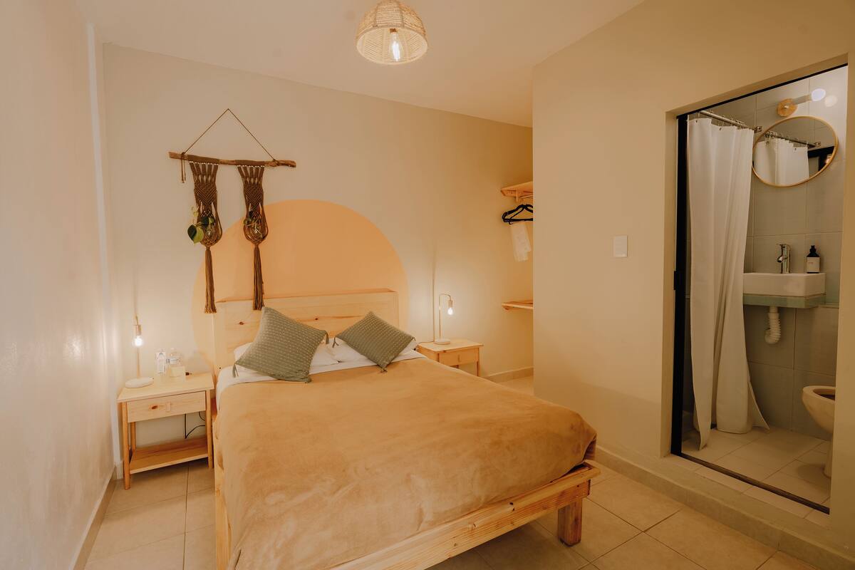Un dormitorio con cama, mesitas de noche, una decoración de macramé colgante, lámpara de noche y un baño con ducha y lavamanos.