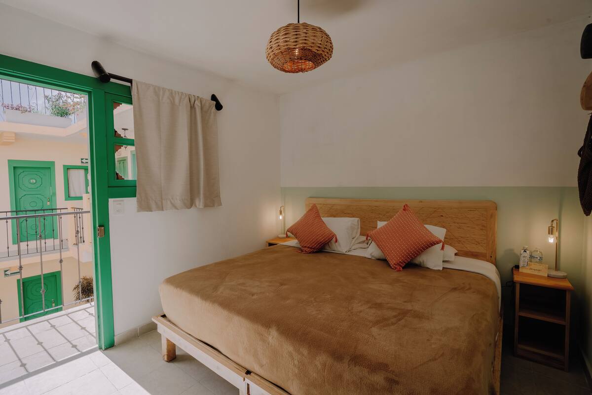 Un dormitorio con cama, mesita de noche, lámpara colgante y vista a un edificio verde en el exterior.