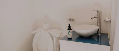 Un baño con un inodoro blanco, un lavabo con encimera azul, un espejo redondo y una pequeña estantería con botellas.