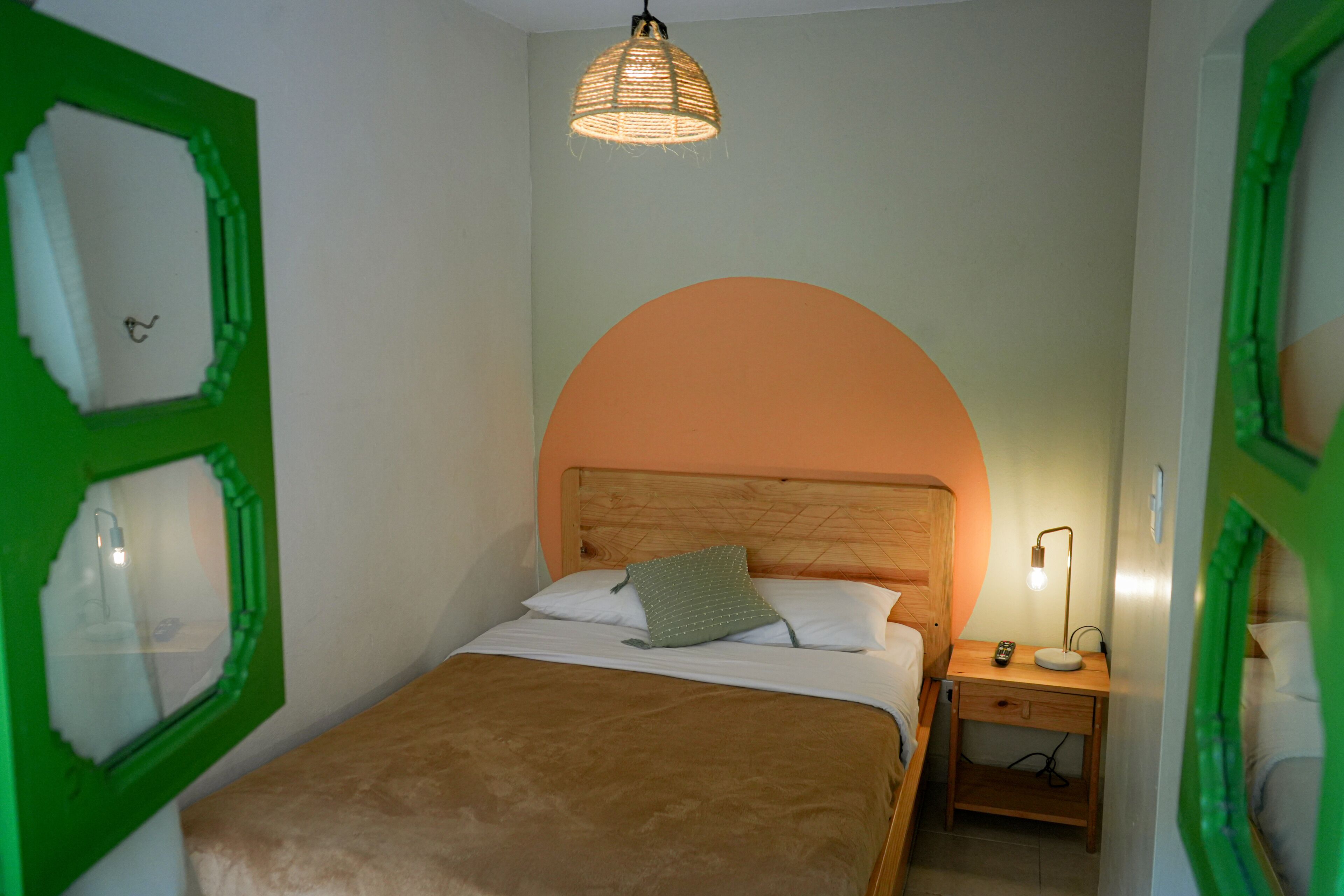 A bedroom with a wooden headboard, a single bed with a brown blanket, a nightstand with a lamp, and a green-framed window.