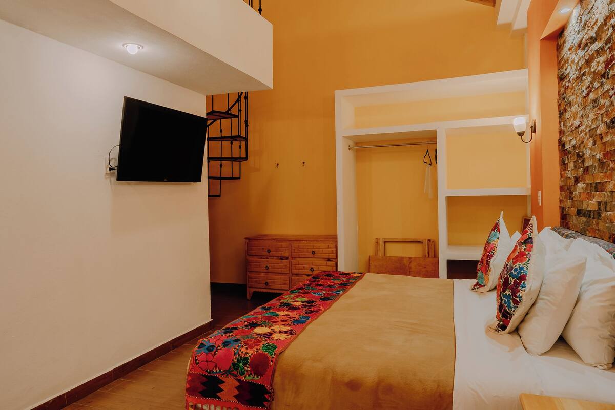 Un dormitorio con techo de madera, una cama con almohadas blancas y de colores, un aparador de madera y un televisor montado en la pared.