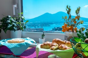 Une table avec des croissants et un gâteau, une plante en pot, et une vue sur une montagne et l’eau.