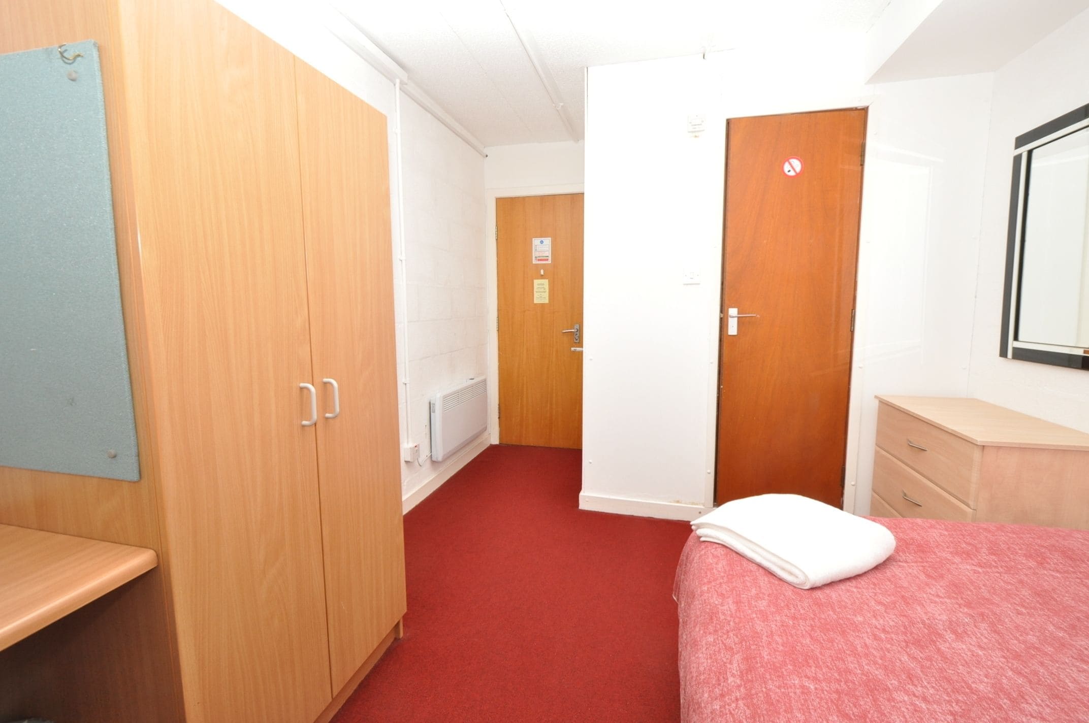 A hotel room with a red carpet, wooden wardrobe, a white bed with a white blanket, a wooden dresser, and a mirror on the wall.