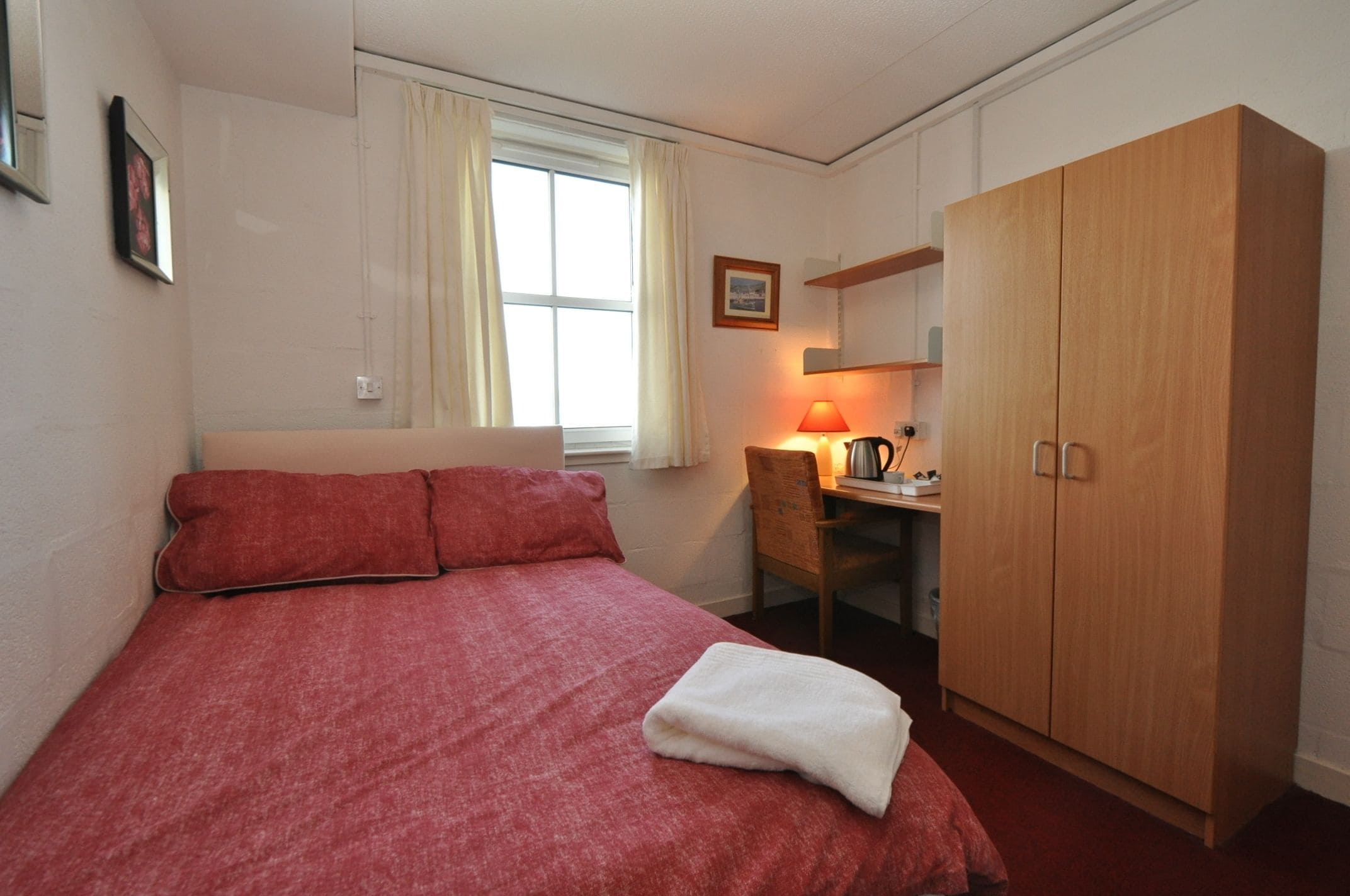 A single-bed room with a red bedspread, a wooden wardrobe, a desk with a lamp, and a window with curtains.