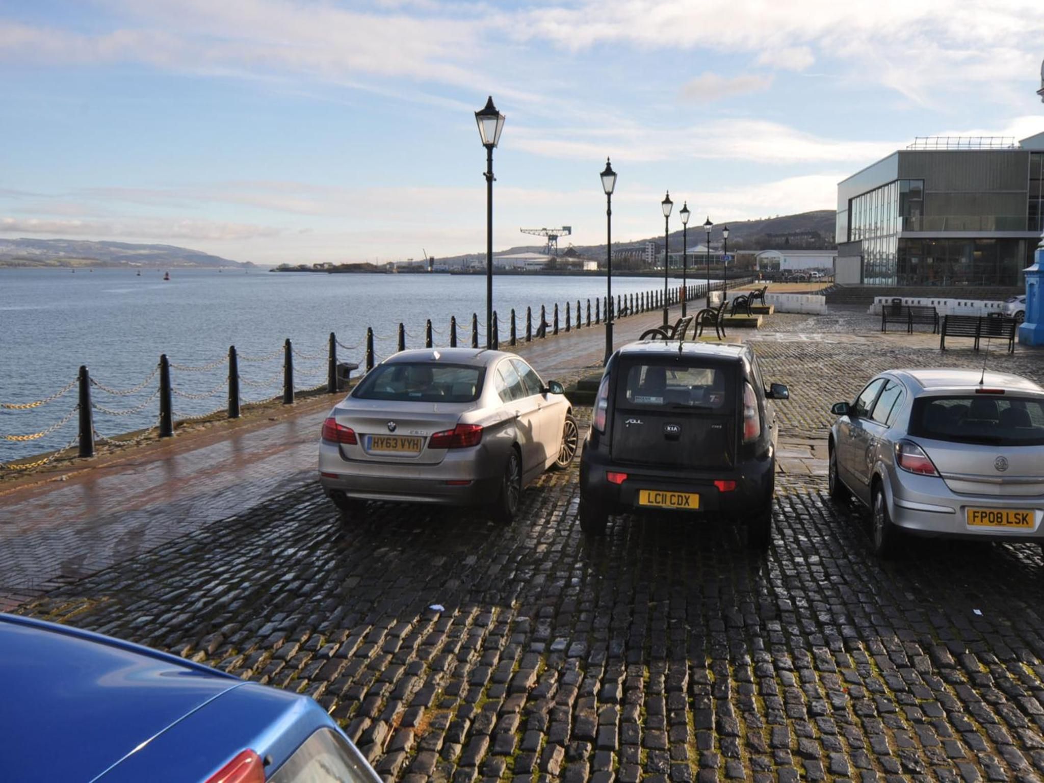 A waterfront parking area with cobblestone pavement and a row of parked cars.