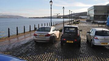 Un espace de stationnement situé au bord de l’eau, avec un revêtement de pavés et une rangée de voitures garées.